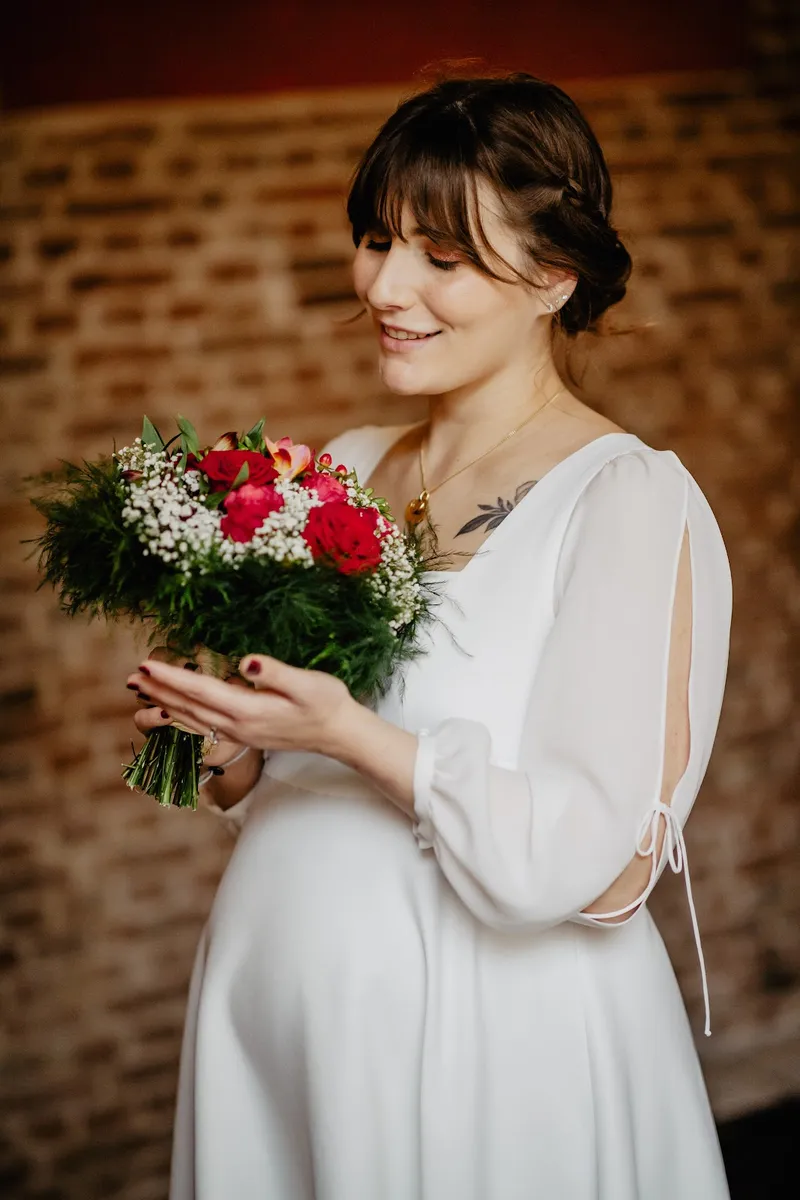 Adelina en robe de mariée sur mesure mi-longue à manches fendues nouées, bouquet de roses rouges et gypsophile à la main