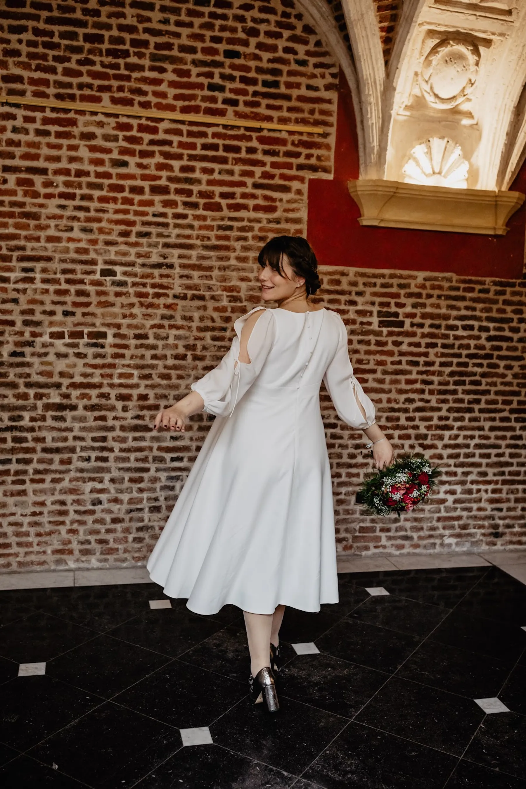 Mariée qui se retourne en souriant, robe blanche évasée mi-longue à manches fendues, sur un sol en damier noir