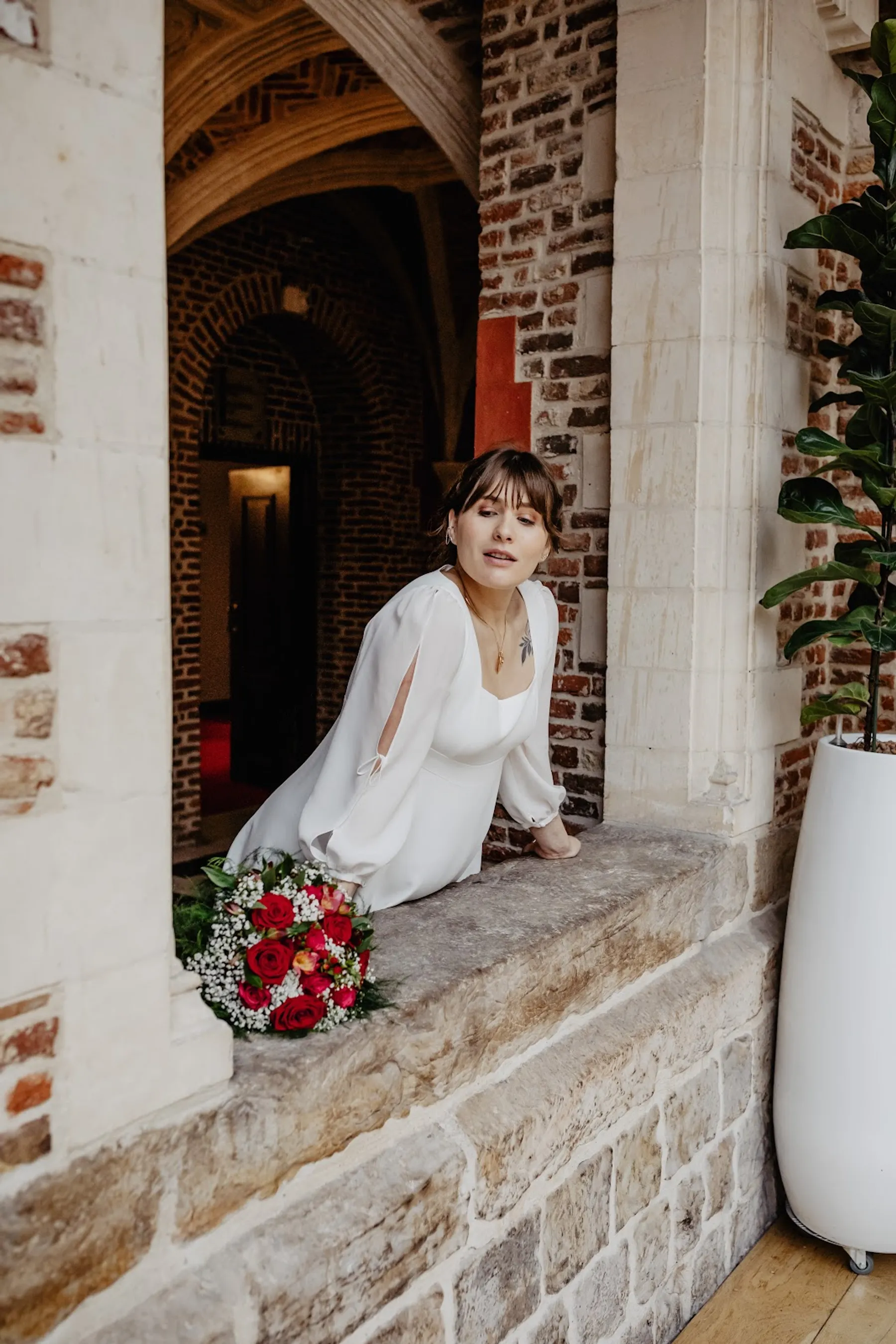 Adelina accoudée au rebord d'une fenêtre en pierre, robe de mariée à manches fendues, bouquet de roses rouges posé devant elle