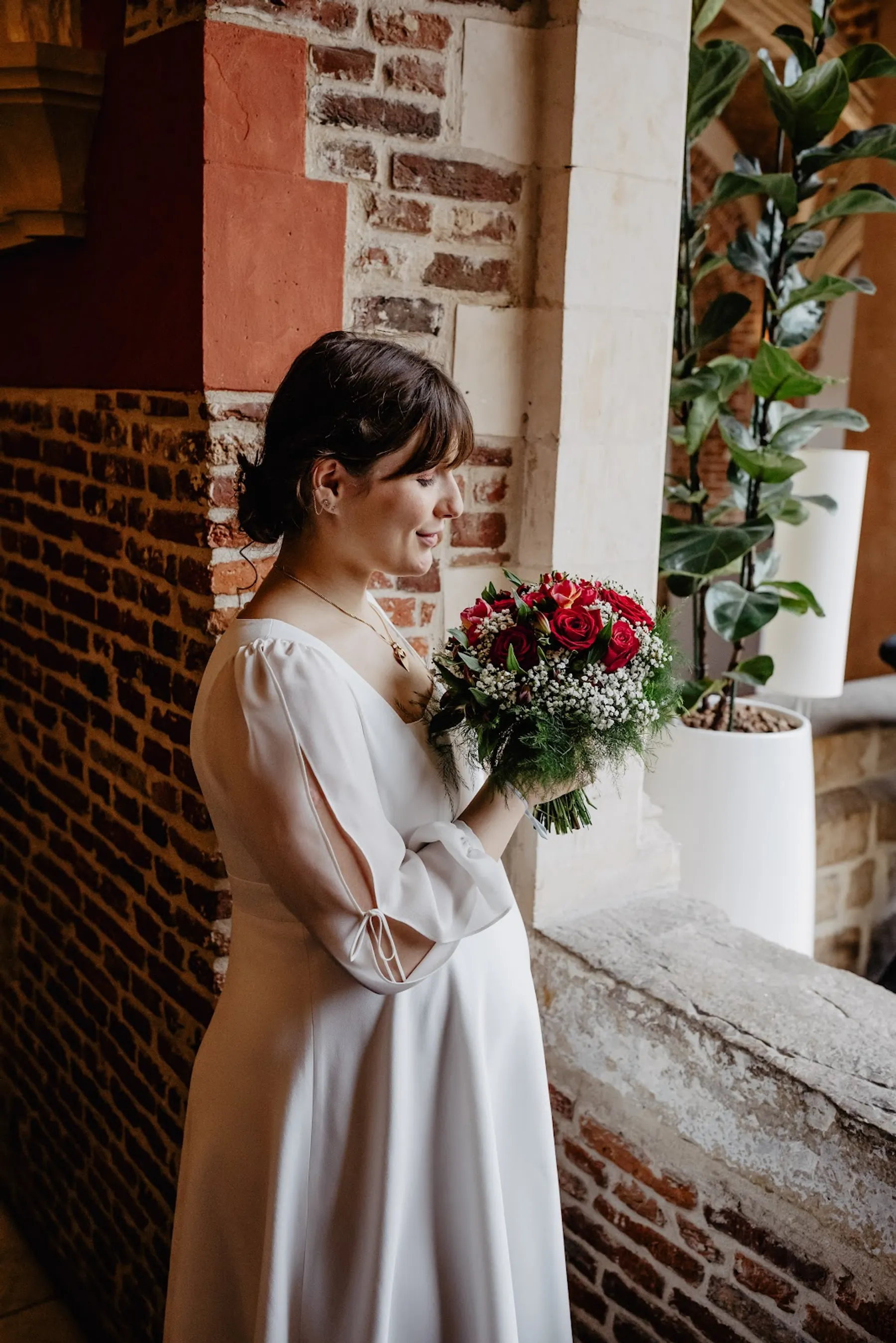 Adelina de profil contre un mur de briques, robe de mariée mi-longue à manches fendues nouées, bouquet de roses rouges