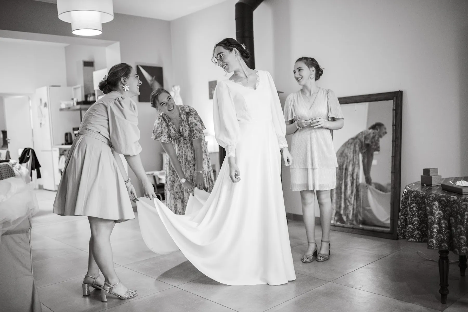 Alice en robe de mariée sur mesure à col V et longues manches, entourée de proches qui soulèvent sa traîne, en noir et blanc