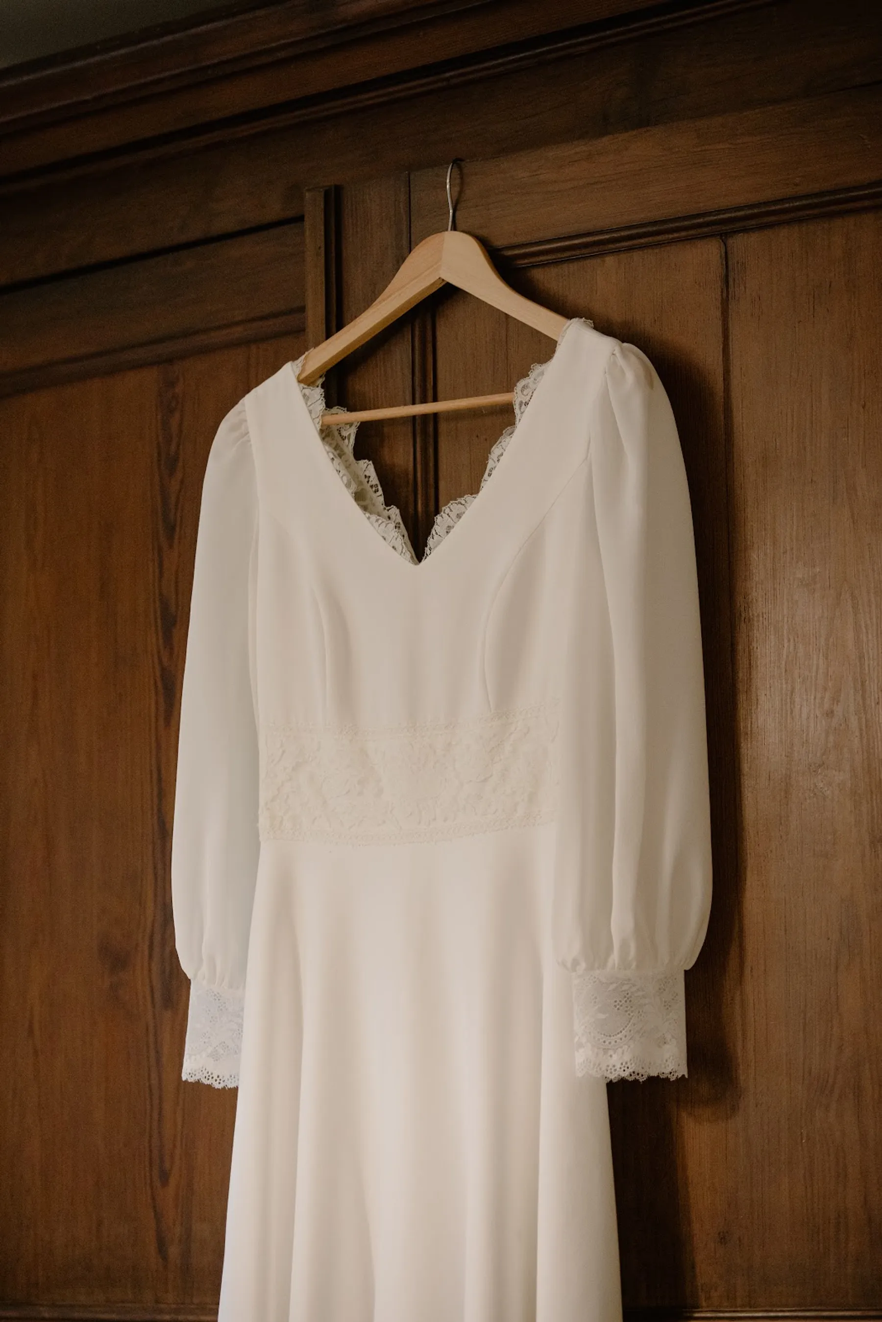Robe de mariée à col V festonné de dentelle et ceinture en dentelle, suspendue à un cintre devant une porte en bois