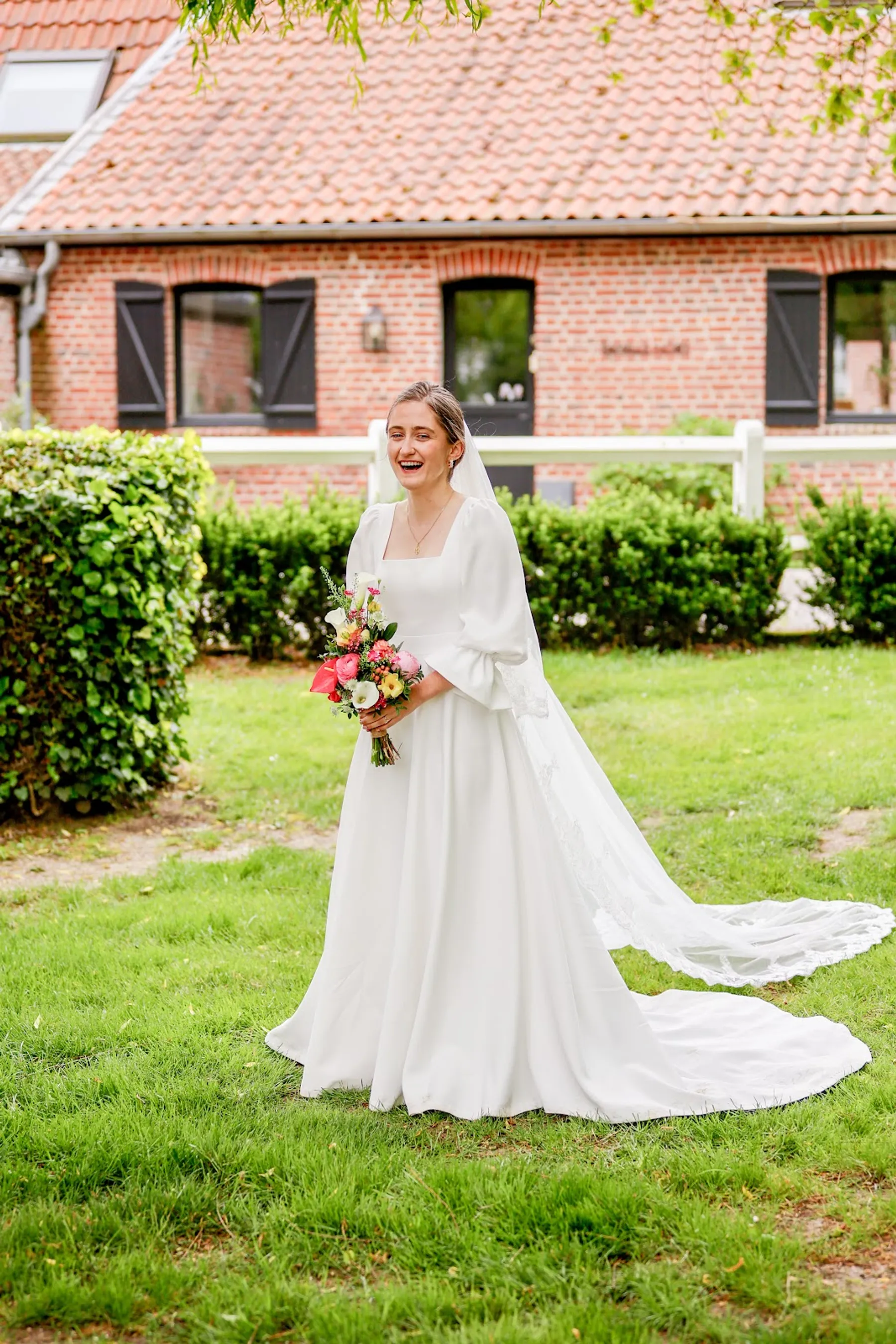 Amélie dans le jardin d'une ferme en briques, robe de mariée à col carré, long voile bordé de dentelle et bouquet coloré