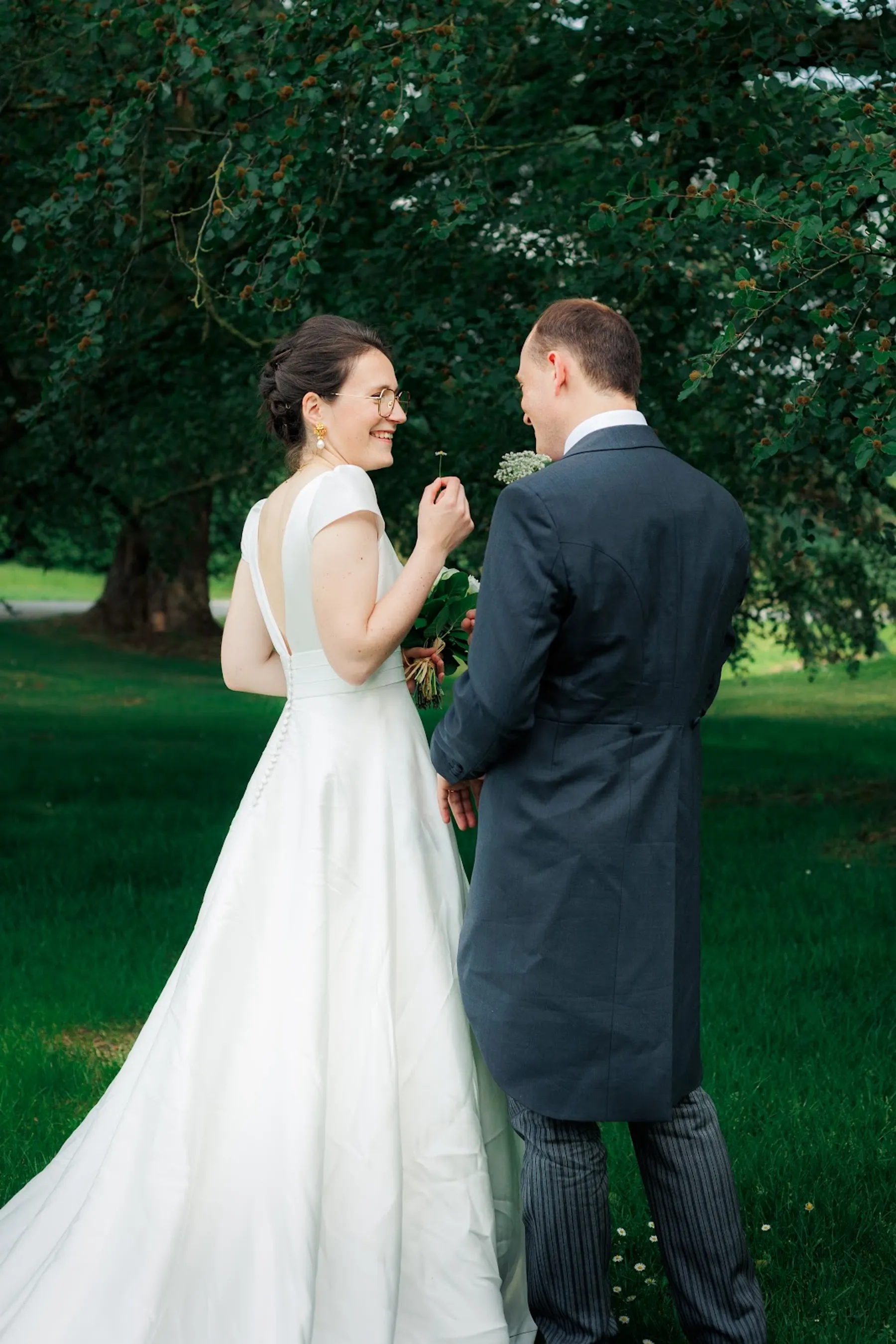 Anne-Laure et son marié de dos dans un parc, robe de mariée à manches courtes et dos échancré, longue traîne en satin