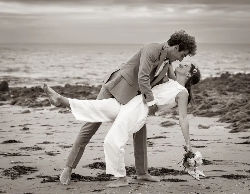 Dorine en combinaison de mariée blanche, cambrée dans les bras de son marié sur une plage rocheuse, en noir et blanc