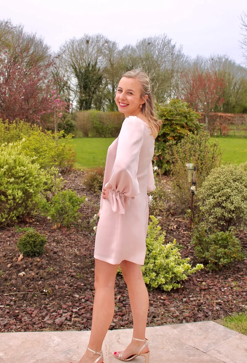 Edmée de dos dans un jardin, robe d'invitée courte rose poudré à manches bouffantes et poignets volantés