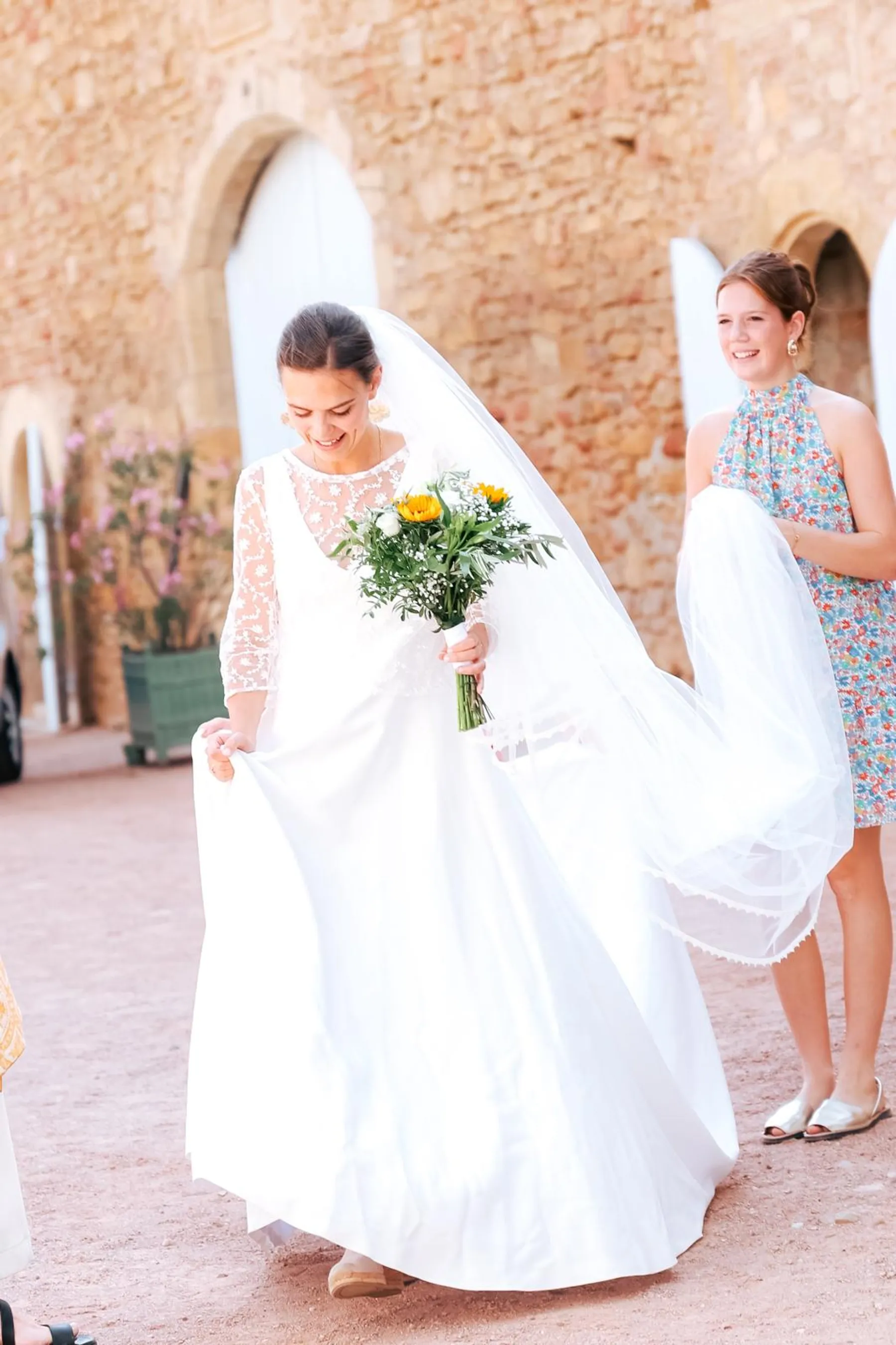 Louise en robe de mariée sur mesure à manches en dentelle, jupe fluide et voile, bouquet de tournesols, soulevant sa jupe
