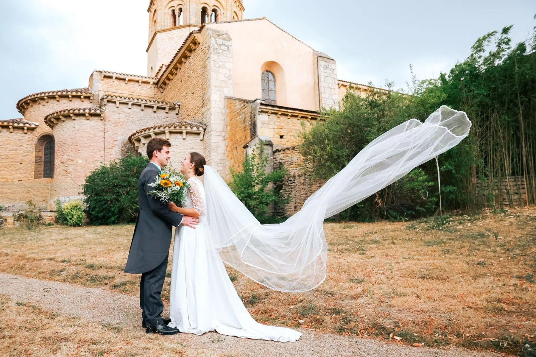 Louise et son marié enlacés devant une église romane en pierre, le long voile de la mariée emporté par le vent