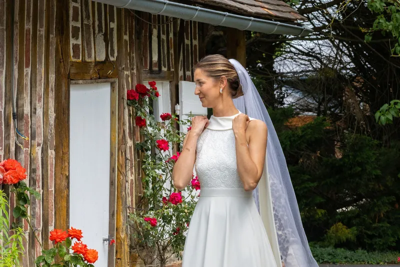 Lucie en robe de mariée sur mesure à col montant sans manches et haut en dentelle, voile long, devant des rosiers rouges