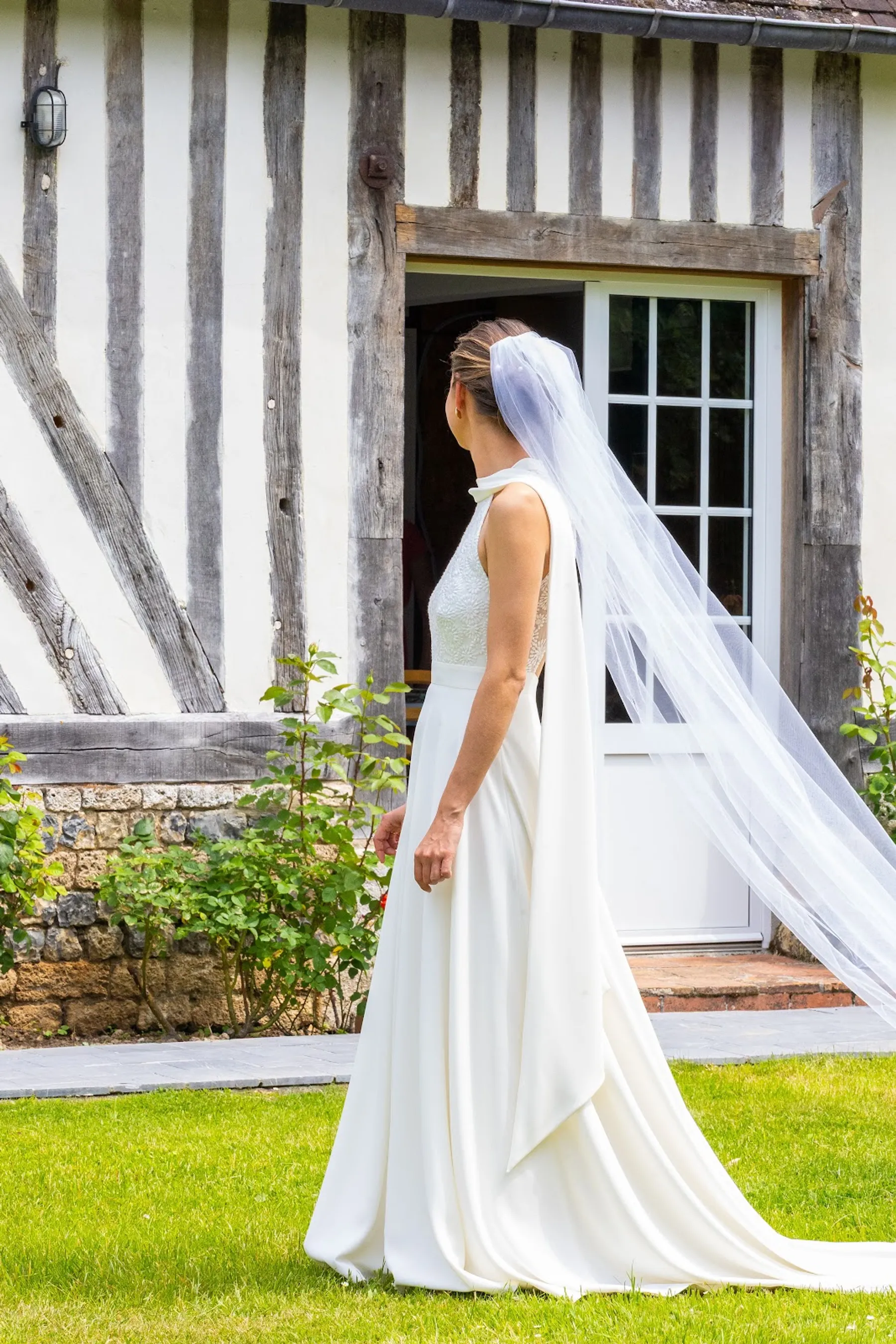 Lucie de dos devant une maison à colombages, robe évasée à col montant avec un drapé dans le dos et un très long voile