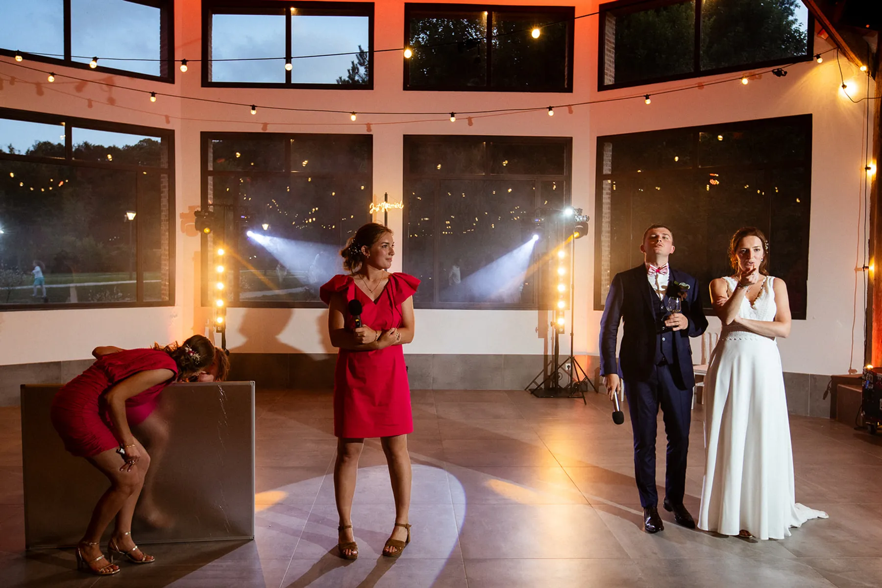 Mathilde en robe rose vif, micro à la main au milieu de la piste sous des guirlandes lumineuses, face aux mariés