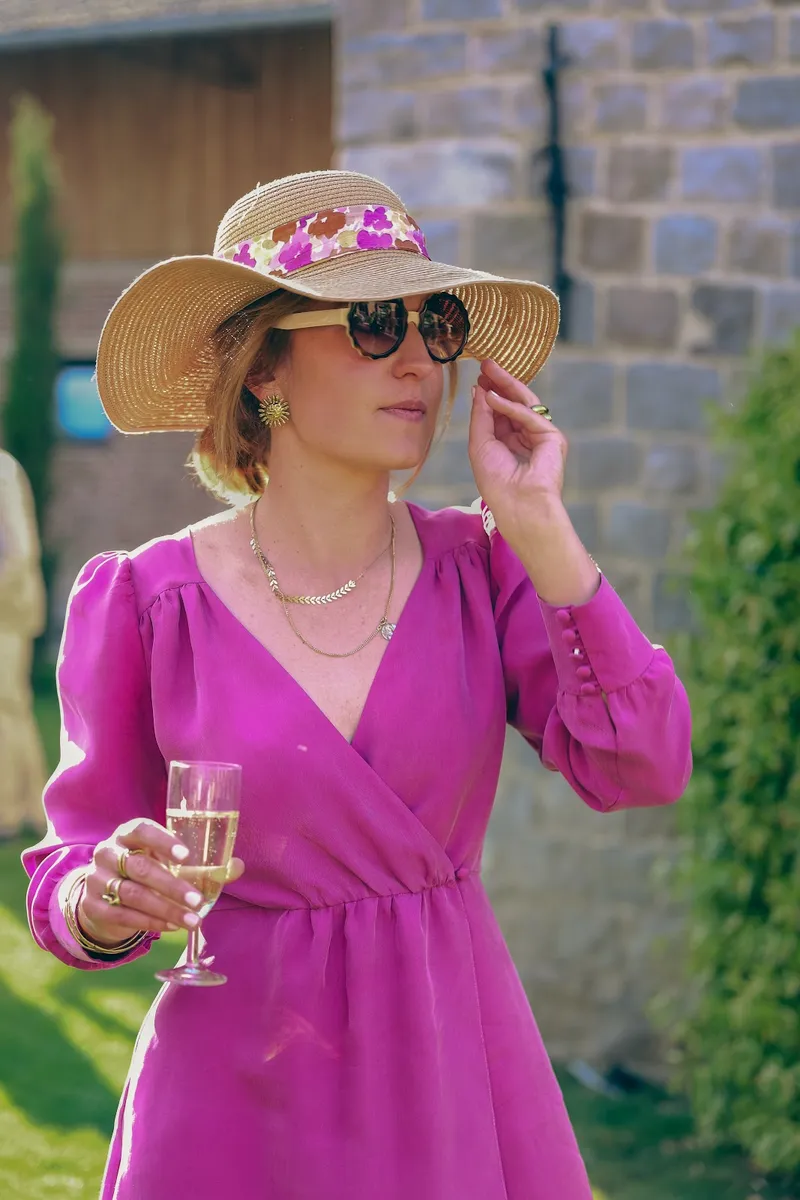 Pauline en robe portefeuille violette à manches bouffantes boutonnées, chapeau de paille à ruban fleuri et lunettes de soleil