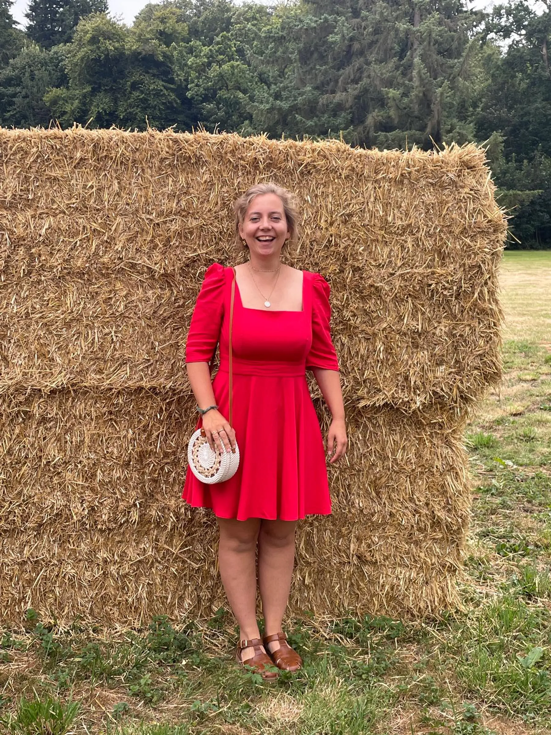 Sarah rit devant une botte de paille, robe rouge à col carré et manches courtes bouffantes, sac en osier rond