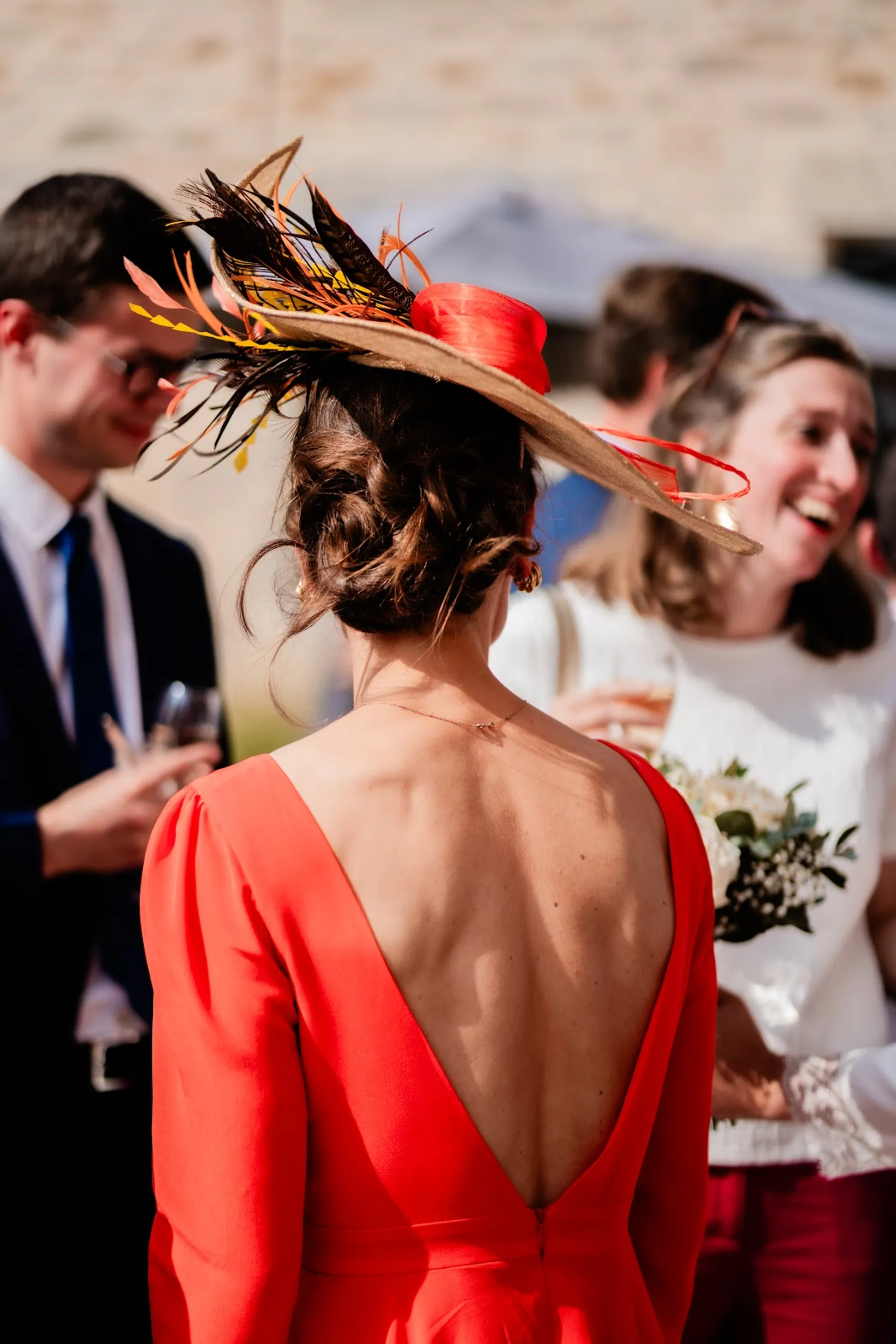 Gros plan sur le dos en V de la robe orangée de Tiphaine et son chapeau à plumes, pendant la réception